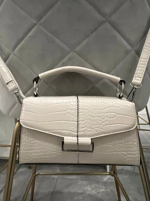 Off-white väska i skinn Primark - Stilren off-white handväska från Primark med krokopräglat mönster och silverfärgade metalldetaljer. Väskan har både handtag och axelrem som går att ta bort, samt ett praktiskt innerfack. Perfekt accessoar för att lyfta din outfit.