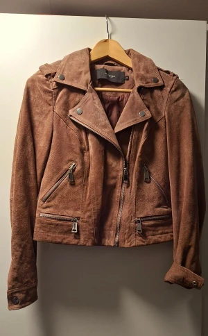 Dimmrosa mockajacka från Vero Moda - Snygg dimmrosa mockajacka från Vero Moda med klassisk bikerstil. Jackan har flera dragkedjor framtill, bred krage med tryckknappar och långa ärmar. Perfekt för dig som vill ha en cool och tidlös look med lite edge.