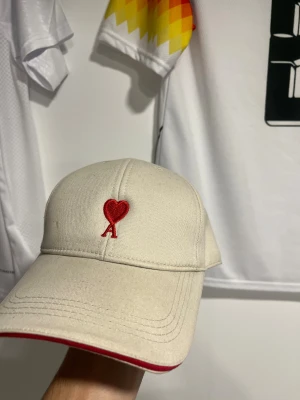 Beige Ami Paris keps - Snygg beige keps från Ami Paris med röd broderad. Tillverkad i bomull och har knappt använts. Den har en minumal fläck lägst framme till vänste (kolla bild 1) men den går troligtvis bort med lite vatten. Använder knappt kepsen därav säljer jag den. 