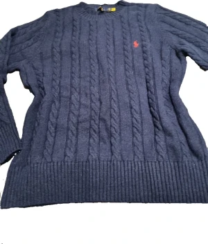 Marinblå kabelstickad tröja Polo Ralph Lauren - Snygg helt oanvänd marinblå kabelstickad tröja med klassisk röd Polo Ralph Lauren logga broderad på bröstet. Tröjan har rund hals, ribbade muddar och lång ärm. Perfekt för dig som gillar stilrent och casual nu inför vintern. 💫 I storlek L fast sitter mer som M. Ny pris: 2395kr