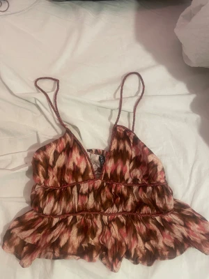 Mönstrad volang topp med tunna band - Säljer en söt topp från H&M med tunna axelband och volangkant. Toppen har ett abstrakt mönster i rosa, brunt och beige och är sydd i ett lätt, luftigt material. Perfekt för varma dagar och ger en bohemisk vibe.
