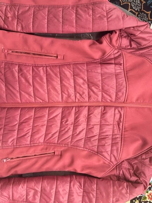 Rosa quiltad pufferjacka från Kayoba - Säljer en snygg rosa pufferjacka från Kayoba Outdoor med quiltade paneler och dragkedjor på fickorna. Jackan har en sportig look och är perfekt för kyliga dagar. Materialet är polyester och jackan har en normal passform.