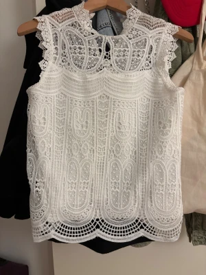 Vit spetsblus med detaljerad mönster - Säljer en vit blus i spets med intricate broderade mönster och snygga kanter. Blusen är ärmlös och har en hög hals med dekorativa spetsdetaljer runt axlar och halsringning. 
