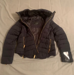 Zara Pufferjacka  - NY OANVÄND MED ETTIKETTEN (se bild)  En supertrendig look, och figurnära jacka med fantastiska guld detaljer. Perfekt för höst, vår och milda vinterdagar! ❄️   Färg: Elegant Marinblå (navy)   Mer detaljer: Hög krage med päls som skyddar mot vind, infällbar huva. Stora, iögonfallande gulddragkedjor fram, på fickorna och som justerbara slitsar på ärmsluten. Fitted passform, mycket fin :) 
