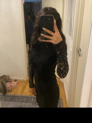 Svart spetsklänning med lång ärm - Säljer en svart kortklänning med långärmade spetsdetaljer och snyggt mönster. Klänningen har en figurnära passform och transparenta ärmar med blommigt spetsmönster. Perfekt för dig som vill ha en elegant look med lite edge.