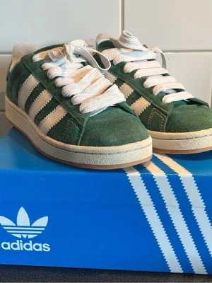 Gröna Adidas Campus 00s sneakers - Säljer ett par gröna Adidas Campus 00s sneakers med klassiska vita ränder och tjocka vita skosnören. Skorna har ovandel i mocka och en beige sula. Perfekta för dig som gillar retrostil och streetwear. Passar bra till både killar och tjejer, bra till våren och sommar! 