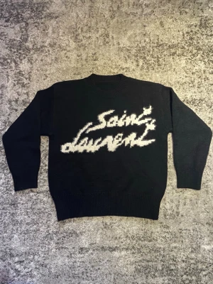 Svart Saint Laurent tröja - Riktigt fet och klassisk Saint Laurent tröja. Grovstickad svart tröja med vit text på, tröjan är i mycket bra skick och storlek S. Hör gärna av dig vid minsta fundering om t.ex. mått, fler bilder etc.😁