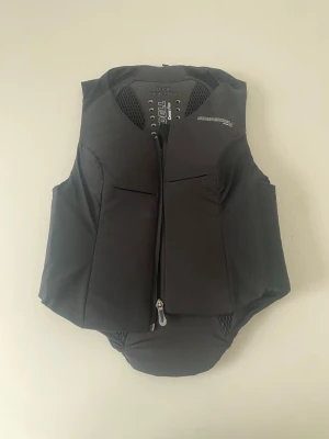 Svart säkerhetsväst från Komperdell - Säljer en svart säkerhetsväst från Komperdell med dragkedja framtill och ventilerande meshpaneler på ryggen. Västen har en sportig design och är tillverkad i slitstarkt syntetmaterial, perfekt för aktiviteter där extra skydd behövs. Säljer då den inte passade när jag fick den. Den är i jättebra skick och endast använd 2 gånger.