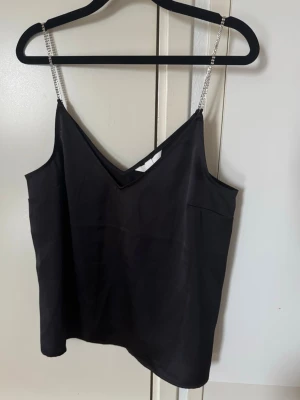 H&M Linne med silvriga axelband stl M - Svart linne från H&M i material i 100% polyester med silvriga axelband, perfekt för utekvällen! Linnet är sparsamt använt och har inga tecken på användning.