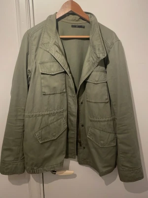 Olivgrön parkajacka med fickor från Åhléns  - Snygg olivgrön parkajacka med fyra stora fickor framtill och klassisk krage. Jackan har långa ärmar och stängs med knappar. Tillverkad i bomull för en bekväm känsla och avslappnad look. Från Åhléns i storlek L. Säljer den för att den sitter för stort på mig, jag e 184cm och 69kg. Inga tecken på defekter. Pris kan sänkas vid snabb affär!