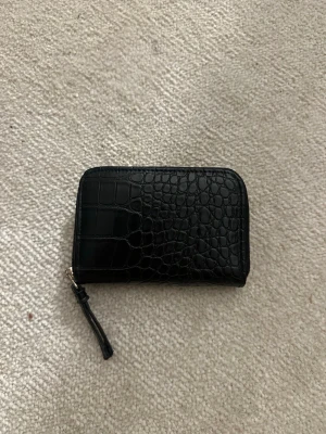 Svart plånbok i krokopräglat skinn - Snygg svart plånbok i krokopräglat skinn med dragkedja runtom och matchande tofs. Perfekt storlek för kort och kontanter, stilren och enkel att ha med sig i väskan. Köpt på åhléns för 250kr men säljer för 90 då jag haft den hemma ett tag men aldrig använt!