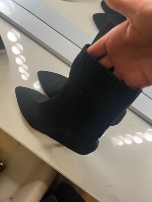 Svarta stretchiga stövletter med klack - Säljer ett par svarta, stretchiga stövletter med spetsig tå och smal klack. De är i ett stickat material som sitter tajt runt ankeln och ger en clean och modern look. Perfekta till både jeans och kjol för en trendig vibe.