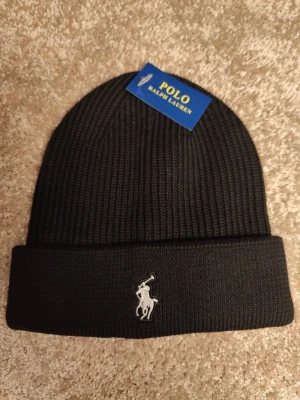 Svart ribbstickad mössa Polo Ralph Lauren - Svart ribbstickad mössa från Polo Ralph Lauren med klassisk uppvikt kant och broderad vit logga framtill. Snygg och enkel design som passar till det mesta. Tillverkad i mjukt material för extra komfort.