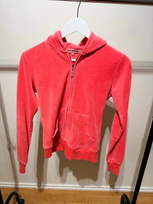 Rosa velourhoodie Juicy Couture - Säljer en ikonisk rosa velourhoodie från Juicy Couture med dragkedja, känguruficka och huva. Baksidan har ett stort guldfärgat Juicy Couture-tryck med krona och detaljer. Perfekt för en avslappnad och trendig look.
