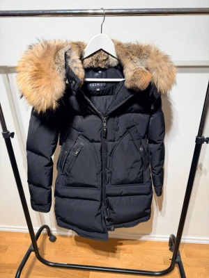 Svart dunjacka med päls från Cedrico - Snygg svart dunjacka från Cedrico med stor fluffig päls runt huvan. Jackan har dragkedja framtill, två snedställda fickor med dragkedja och quiltad design. Perfekt för kalla vinterdagar och ger en riktigt lyxig vibe jackan passar dig som är mellan 150cm till 160cm och upp till 60kg. Jackan är knappt använd och köpt för 8500.perfekt jacka på vintern för en lyxig känsla.