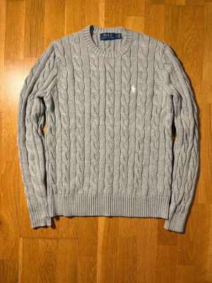 Grå kabelstickad tröja Polo Ralph Lauren - Snygg grå kabelstickad tröja från Polo Ralph Lauren med rund halsringning och klassisk vit broderad logga på bröstet. Tröjan har ribbade muddar vid ärmslut och nederkant. Perfekt för dig som gillar stilren och tidlös design.