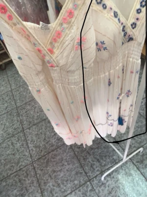 Vit långklänning med broderade blommor - Vit klänning med v-ringning och broderade blommor i rosa och blått. Klänningen har smock i midjan, tunna axelband och volangdetaljer. Köpt i Spanien. Aldrig använd💗