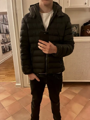 Svart pufferjacka från Colmar - Snygg svart pufferjacka från Colmar med quiltad design och huva. Jackan har dragkedja framtill och två sidofickor med dragkedja. Perfekt för kyliga dagar och ger en modern, sportig look.