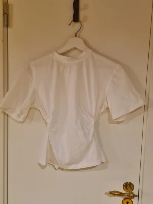 Vit omlottopp med kort ärm - Snygg vit topp med omlottdetalj i midjan och korta ärmar. Toppen har en figurnära passform och är tillverkad i ett mjukt material som känns bekvämt mot huden. Perfekt att styla med jeans eller kjol för en clean look.