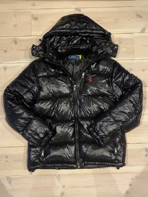 Svart dunjacka från Polo Ralph Lauren - HELT NY/OANVÄND!! Snygg svart dunjacka från Polo Ralph Lauren med huva och dragkedja framtill. Jackan har quiltad design, två fickor med dragkedja och den klassiska röda loggan på bröstet. Perfekt för kalla dagar och riktigt stilren look. Först till kvarn!!