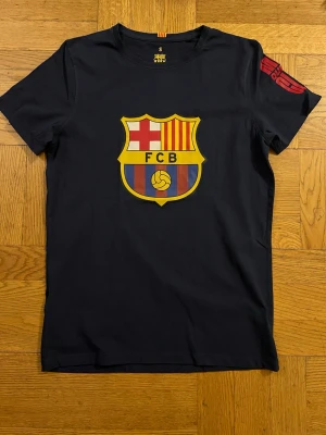 FC Barcelona marinblå t-shirt - Snygg marinblå t-shirt från FC Barcelona med stort klubbmärke på bröstet och röda tryck på ärmen och ryggen. Klassisk passform och korta ärmar, perfekt för dig som älskar fotboll och vill visa ditt lagstöd. Tillverkad i mjuk bomull för skön känsla.