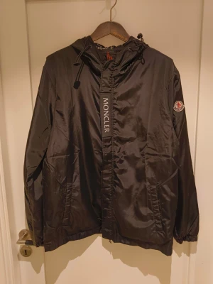 Svart vindjacka från Moncler - Snygg svart vindjacka från Moncler med huva och dragsko. Jackan har dragkedja framtill, två fickor och Moncler-logga på ärmen. Insidan av huvan har ett mönster med Moncler-tryck i vitt och rött. Perfekt för dig som vill ha en stilren och sportig look. Fodret trasigt på insidan vid högra armhålan, inget som påverkar utseendet nämnvärt.