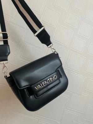 Svart axelväska från Valentino - Stilren svart axelväska från Valentino med bred, justerbar axelrem i svart och vitt. Väskan har ett elegant metallogo framtill och är tillverkad i slätt skinnliknande material. Perfekt för att lyfta din outfit med en trendig touch.