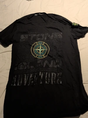 Svart  t-shirt med tryck - Svart t-shirt med stort tryck på bröstet där det står 'S.I Adventure' och den klassiska kompassloggan i mitten. T-shirten har rund hals och korta ärmar, samt en patch med loggan på ärmen. Perfekt för dig som gillar streetwear. Köpt här, så osäker på om den är äkta. 
