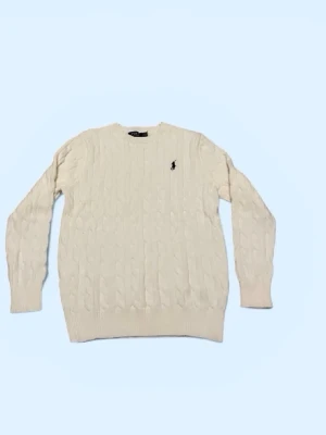 Vit kabelstickad tröja Polo Ralph Lauren - Kabelstickad vit tröja från Polo Ralph Lauren med rund halsringning och den klassiska broderade loggan på bröstet. Tröjan har långa ärmar och ribbade muddar vid ärmslut och nederkant. Perfekt för dig som gillar stilrena och tidlösa plagg.