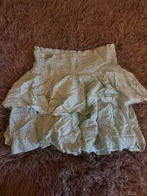 Grön volangkjol med blommigt mönster - Söt kortkjol i ljusgrönt med små vita blommor och bred smock i midjan. Kjolen har flera lager volanger som ger en fluffig och lekfull look. 