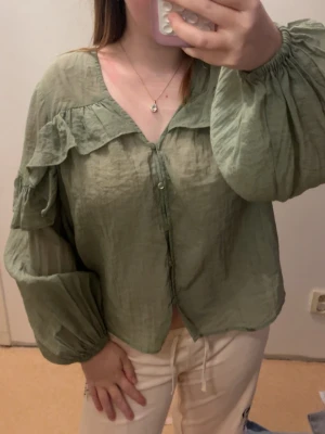 Grön volangblus med puffärm - Snygg ljusgrön blus med volangdetaljer över axlarna och knäppning framtill. Blusen har ballongärmar med resår vid handlederna och är tillverkad i ett tunt, lätt och något transparent tyg som ger en luftig känsla. Perfekt för en trendig och avslappnad look.