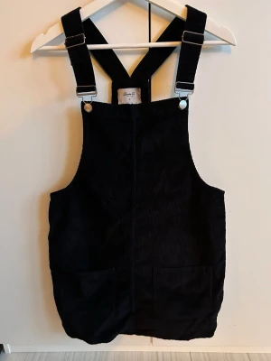 Svart manchesterbyxdress med fickor - Snygg svart byxdress i manchester med breda axelband som korsas i ryggen och stora fickor framtill. Perfekt för dig som gillar en avslappnad och trendig stil. Justerbara spännen på axelbanden för bästa passform.