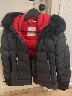 Moncler svart dunjacka med röd insida - Snygg svart dunjacka från Moncler med röd insida och huva med svart fuskpäls. Jackan har två dragkedjefickor framtill och quiltad design. Perfekt för sveriges kalla vinter. köpt några år sen men knappt använd.PRIS KAN DISKUTERAS