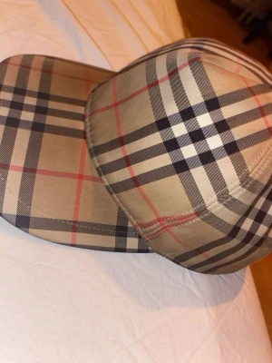 Burberry keps beige  - Snygg keps från Burberry i deras ikoniska beigea rutmönster med svarta, vita och röda linjer. Tillverkad i bomull och polyester, perfekt för att lyfta din streetwear-look. Märkeslapp på insidan och justerbar passform.