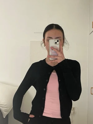 Svart basic kofta med rund hals - En svart, tunn och enkel kofta med rund halsringning och knäppning framtill. Perfekt att slänga över en t-shirt eller linne. Långärmad och figurnära modell som passar till det mesta.