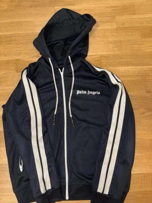 Palm Angels svart loungewear set - Snyggt svart loungewear set från Palm Angels med vita ränder längs ärmar och ben. Setet består av en hoodie med dragkedja och matchande byxor, båda med Palm Angels-logga i vitt. Tillverkad i mjukt och stretchigt material, perfekt för chill dagar hemma.