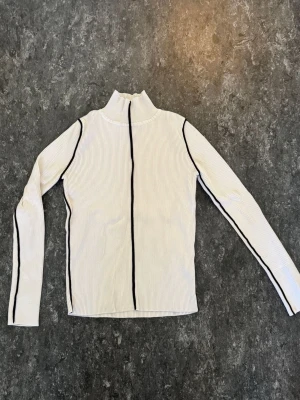 Vit ribbad polotopp med svarta sömmar - Säljer en vit ribbad polotopp med svarta kontrastsömmar längs ärmar och framtill. Toppen har lång ärm och hög krage, perfekt för en clean och edgy look. Passar till både jeans och kjol för en trendig stil.