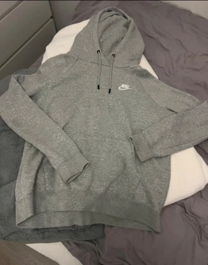 Grå hoodie från Nike - Klassisk grå hoodie från Nike med vit logga på bröstet. Hoodien har dragsko i huvan och en stor magficka. Perfekt för en avslappnad stil och enkel att matcha med andra plagg.
