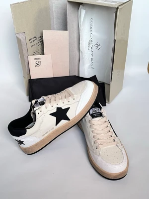 Golden Goose sneakers med svart stjärna - Snygga sneakers från Golden Goose i vitt och beige skinn med svart stjärndetalj på sidan och svart häl. Klassisk rund tå, platt sula och snörning framtill. Perfekta för en trendig och avslappnad stil.