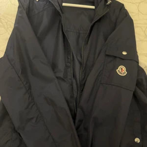 Marinblå vindjacka från Moncler - Snygg marinblå vindjacka från Moncler med dragkedja och ficka på ärmen med logotyp. Jackan har huva och stilrena detaljer i silver. Perfekt för blåsiga dagar och enkel att matcha med allt. Storlek 5 passar L/XL. Vid frågor kontakta mig! 