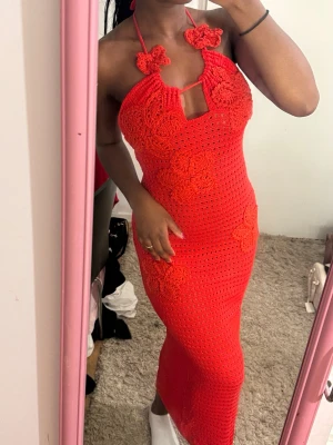 Röd virkad långklänning med blommor - Trendig röd långklänning i virkad stil med blommiga detaljer och halterneck. Klänningen har öppen rygg och figurnära passform, perfekt för sommarens alla tillfällen. Snygg cut-out framtill och knyt i nacken.