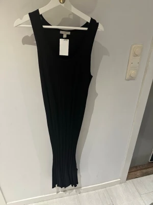 Svart ribbad långklänning  - Stilren svart långklänning från H&M med ribbad struktur. Klänningen är ärmlös och har en rund halsringning. Materialet är mjukt och stretchigt, vilket ger en smickrande passform. Perfekt för dig som gillar minimalistisk och modern stil.