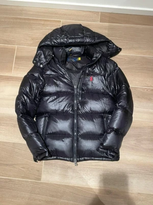Ralph Lauren Svart Puffer Jacka  - Snygg svart pufferjacka från Polo Ralph Lauren med huva och dragkedja framtill. Jackan har det klassiska röda Polo-logotypen broderad på bröstet och två sidofickor med dragkedja. Perfekt för kalla dagar och riktigt trendig look.