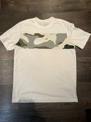 Nike - Nike Dri-Fit t-shirt i vitt med en bred camouflagemönstrad panel över bröstet. Klassisk rund hals och korta ärmar. Tillverkad i ett lätt och funktionellt material som transporterar bort fukt, perfekt för en aktiv livsstil.