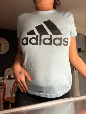 Ljusblå Adidas t-shirt med logga - Snygg ljusblå t-shirt från Adidas med stor svart logga framtill. Klassisk rund hals och korta ärmar. Perfekt för en sportig och avslappnad stil. Tillverkad i mjuk bomull för skön komfort.