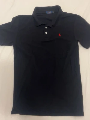 Svart pikétröja från Polo Ralph Lauren - Klassisk svart pikétröja från Polo Ralph Lauren med röd broderad logga på bröstet. Tröjan har korta ärmar, krage och knäppning med två vita knappar. Perfekt till jeans eller shorts för en clean och stilren look. Storlek m/s