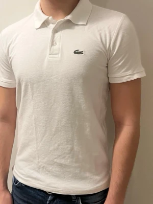 Vit pikétröja från Lacoste - Klassisk vit pikétröja från Lacoste med korta ärmar och krage. Tröjan har knappar framtill och den ikoniska krokodilloggan broderad på bröstet. Perfekt till jeans eller shorts för en clean och stilren look.storlek m/s