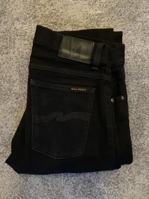 Nudie Jeans - Snygga svarta jeans från Nudie Jeans med klassisk femficksdesign och diskret logga på bakfickan. Jeansen har smal passform och är tillverkade i mjukt denimtyg. Perfekta för dig som gillar stilrena och tidlösa plagg.