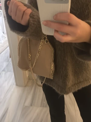 Beige handväska från Stella McCartney - Trendig beige handväska från Stella McCartney med kedjedetaljer i guld och rund logotypbricka. Väskan har en mjuk struktur och kedjehandtag som ger en cool och modern look. Perfekt accessoar för att lyfta din outfit👏👏💓🥰🥰🥰
