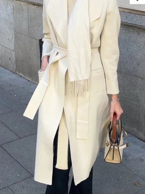 Vit omlottkappa med bälte - Stilren vit kappa och brett bälte i midjan. Kappan har stora slag, långa ärmar och två fickor framtill. Perfekt för dig som gillar en clean look